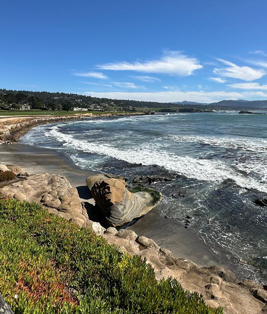 Scenic coastal view with waves crashing against rocky shore under a bright blue sky.